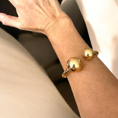 Bangle de acero dorado con diseño abierto y esferas en los extremos. Minimalista y elegante.